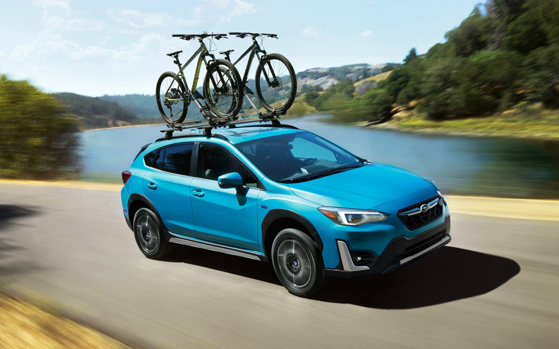 A blue Crosstrek Hybrid with two bicycles on its roof rack driving beside a river | Dutch Miller Subaru in Charleston WV